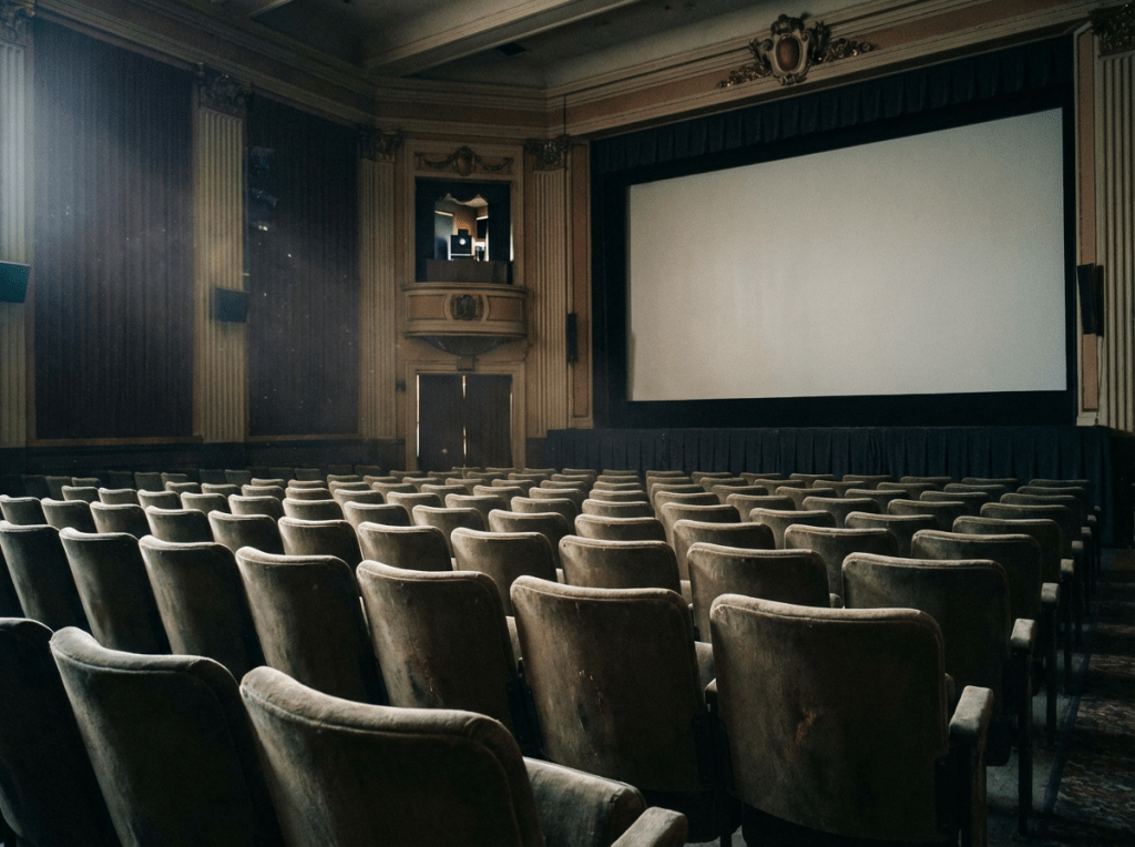 Rows of velvet seats in an ornate, empty vintage movie theater facing a large blank screen.